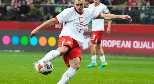 Piłkarz w białej koszulce z orłem i czerwonych spodenkach kopie piłkę na boisku.